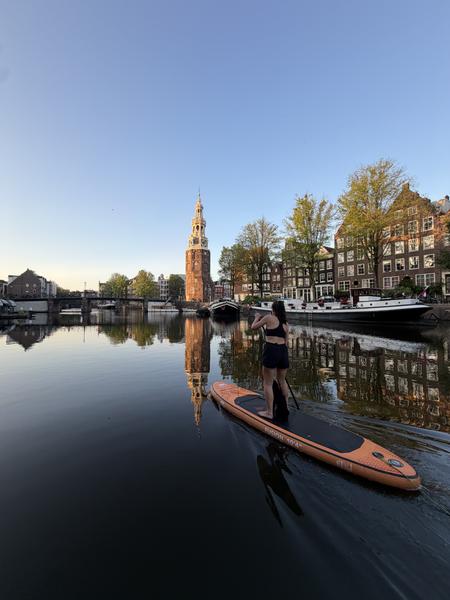 A person enjoys stand-up paddleboarding on a calm canal in Binnenkant, Centrum, Amsterdam, under a clear morning sky